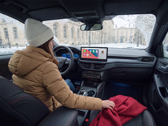 Frau im Auto bei Schnee, trägt eine Mütze und eine Jacke, mit einem Bildschirm in der Mitte und einer Wolldecke auf den Knien.